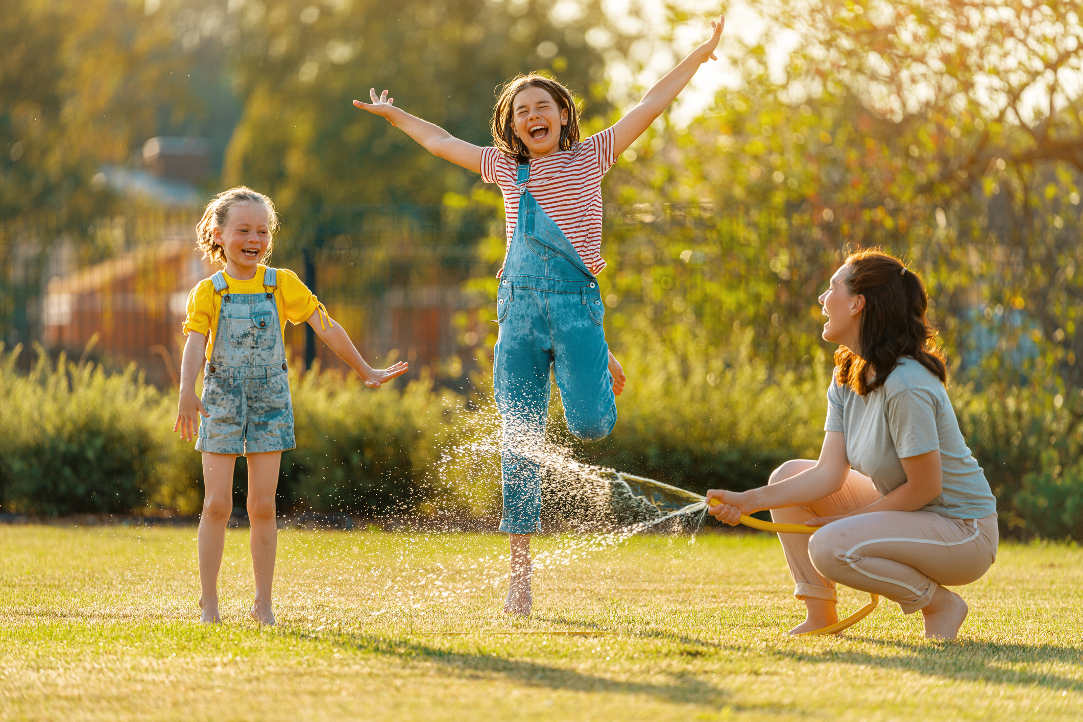 Happy family playing in backyard. Mother sprinkling her kids in hot summer day.