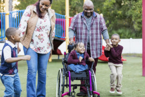 A happy African-American family with three young children walking together on a playground. The daughter, 5 years old, is in a wheelchair. She has caudal regression syndrome, a rare congenital disorder which affects the development of the lower spine. She is in the middle between the parents. Her brothers, twin 4 year old boys, are on the ends, holding their parents' hands.