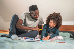 Smiling African-American single father having fun, coloring together with his little son in the family bedroom. Low angle shot, full length image.