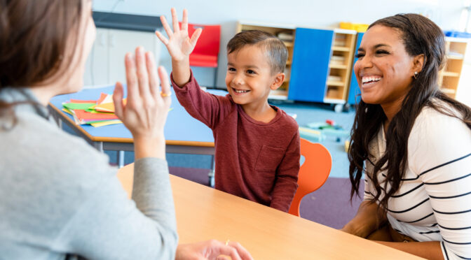 A cheerful teacher gives a little boy a high five during a parent-teacher conference. The boy's mom smiles cheerfully.