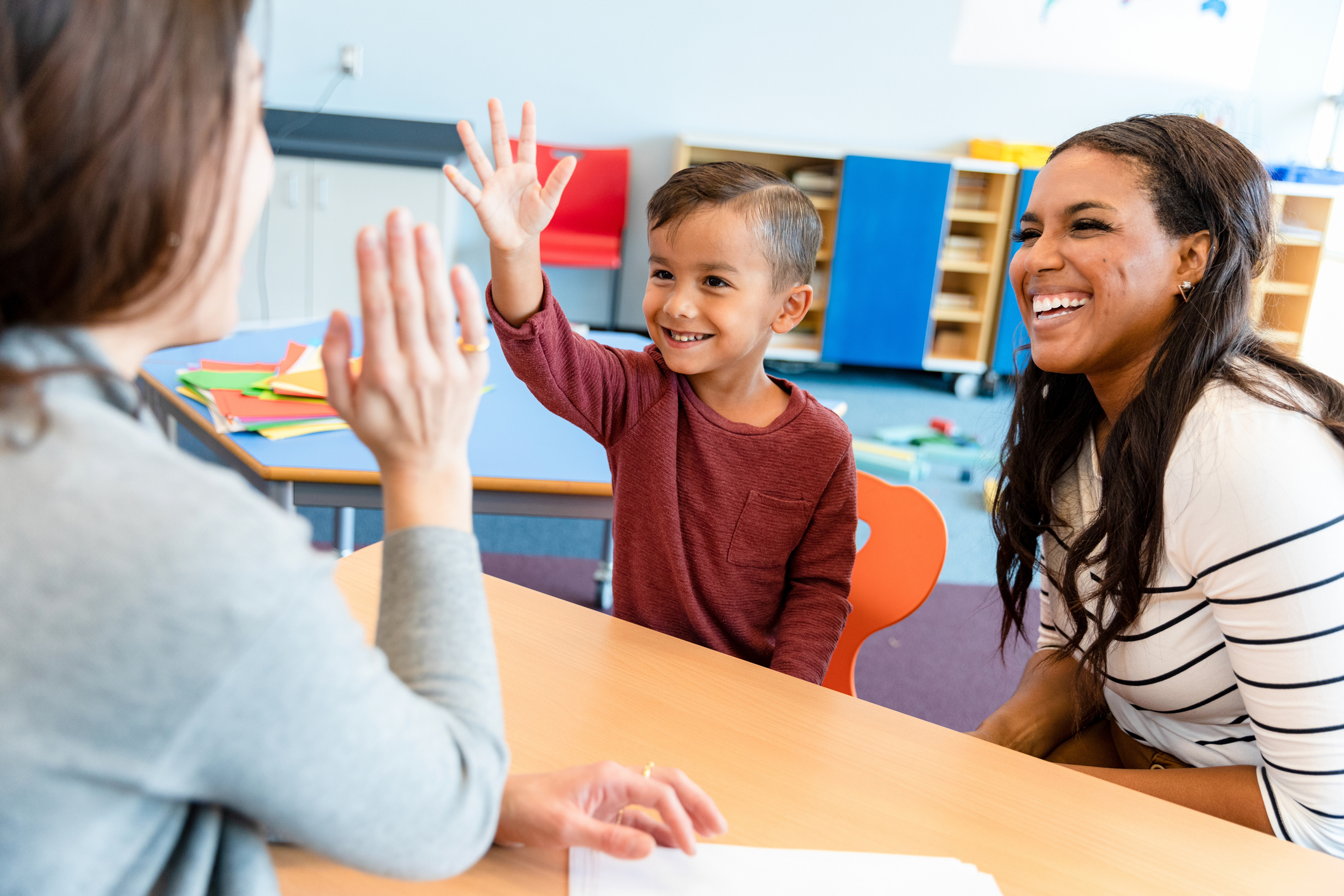 A cheerful teacher gives a little boy a high five during a parent-teacher conference. The boy's mom smiles cheerfully.