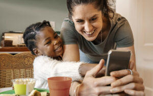Mother and adopted daughter looking at a mobile phone, smiling at the dining table in the kitchen at home