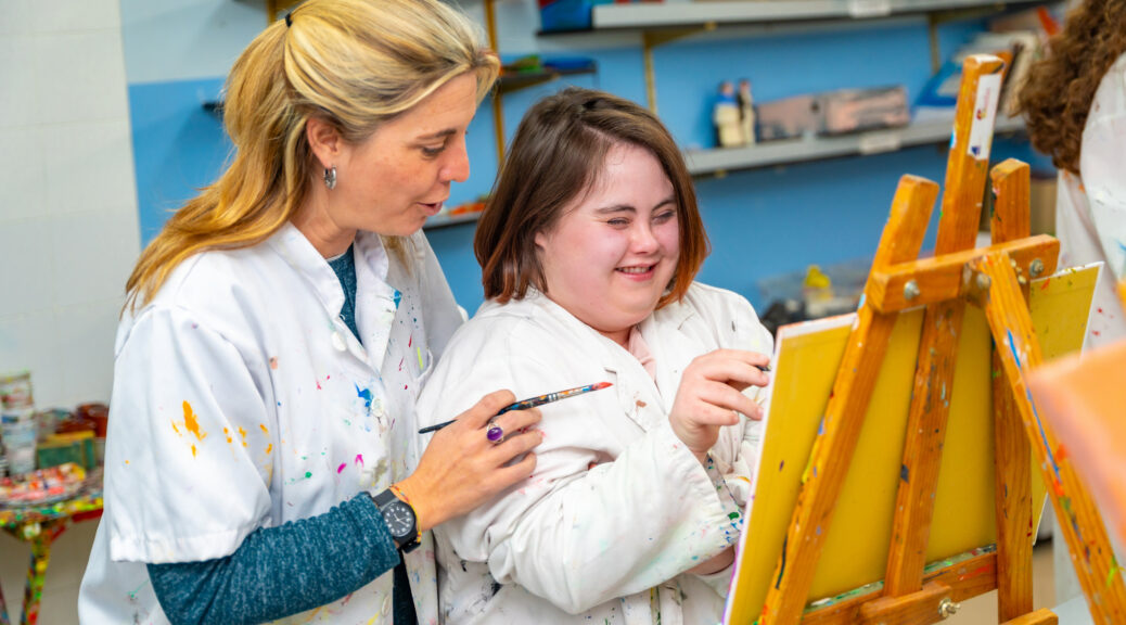 Adult caucasian teacher helping a smiling woman with down syndrome during painting class