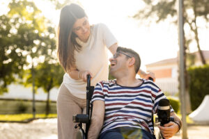 An adult man with a disability sits in an automatic wheelchair with a young woman in a park at sunset.The woman and the man look at each other happily. 