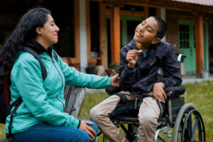 Disabled hispanic child is sitting in his wheelchair, holding a flower and smelling it, while his mother sits next to him and looks at him with love