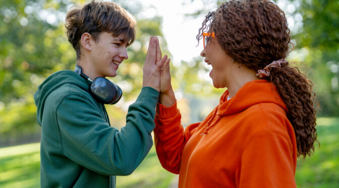 Two friends stand in a park playing a game of high five. The sun shines down as they laugh and celebrate their friendship in a lively outdoor setting.