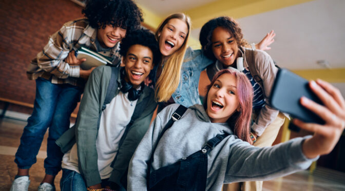Group of happy high school students having fun while taking selfie with cell phone in hallway.
